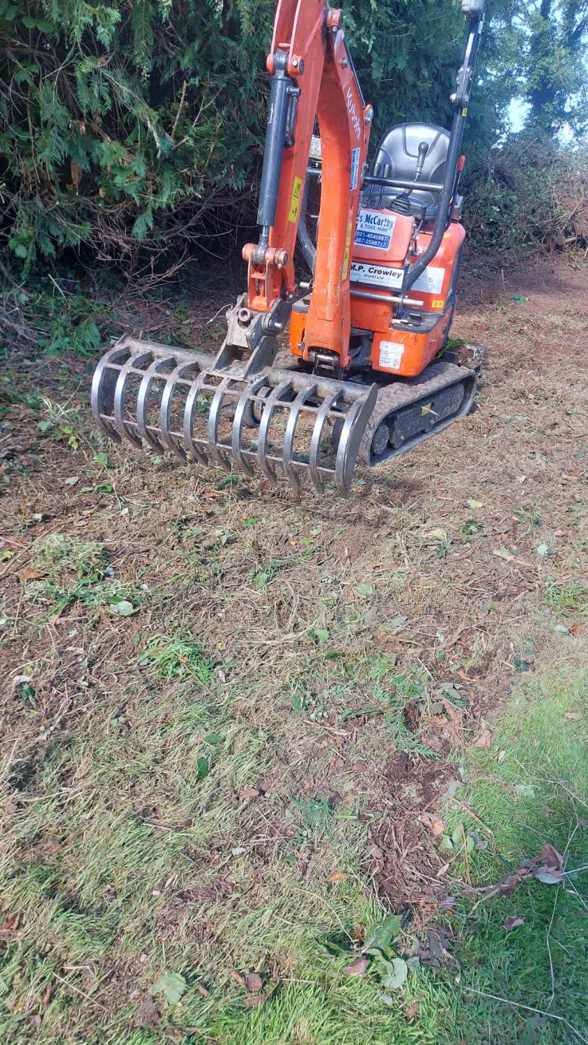 Mini digger with a rake attachment clearing ground beside a tree line